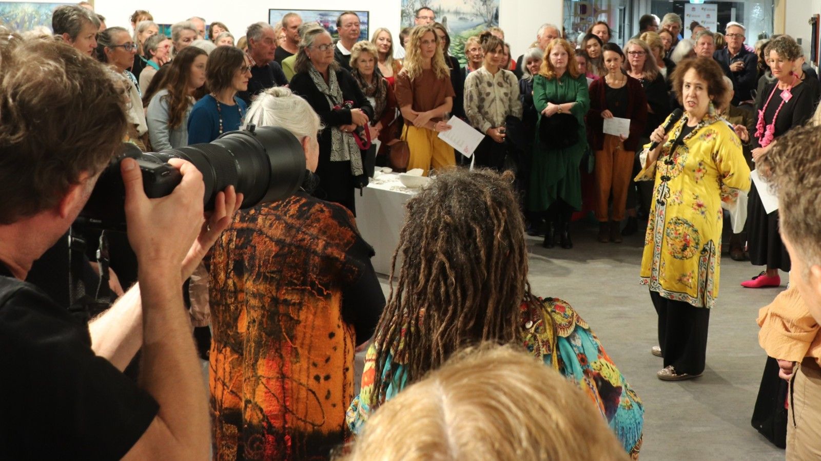 A woman dressed in a stunning yellow coat speaking to an audience of art lovers. banner image