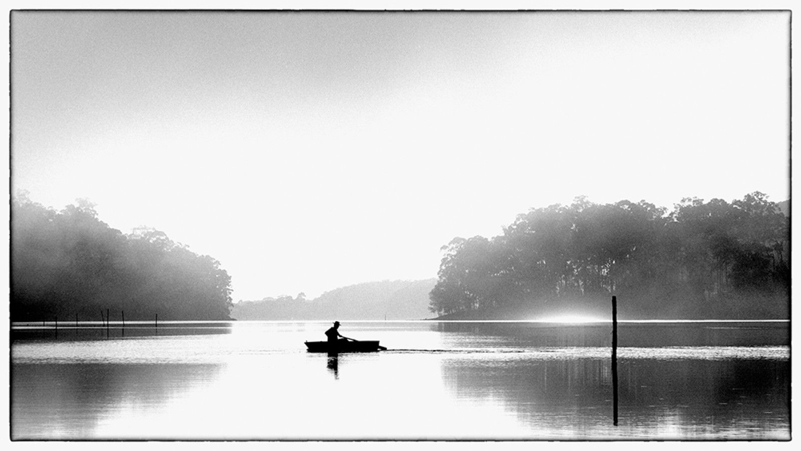 A black and white photo of a man out on a lake in a boat. banner image